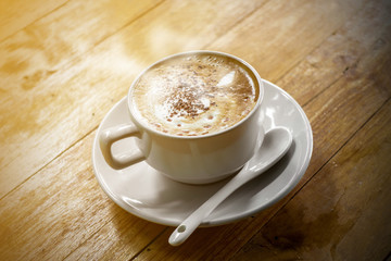 The  white ceramic cup of hot capuccino coffee on the wooden table in the restaurant or coffee shop with flare in the natural light. 