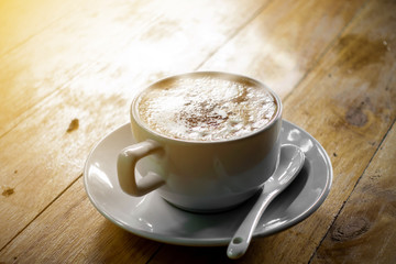 The  white ceramic cup of hot capuccino coffee on the wooden table in the restaurant or coffee shop with flare in the natural light. 