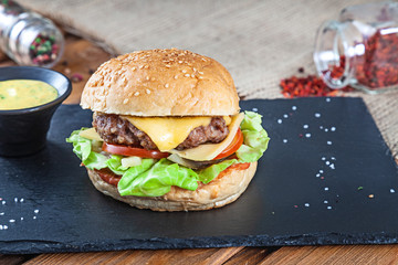 Fresh tasty burger with cheese, lettuce, tomato, cucumber on black stone with sauce. American fast food. Cheeseburger with copy space on wooden background. Close up, selective focus. Food. Grill menu