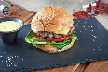 Fresh tasty burger with cheese, lettuce, tomato, cucumber on black stone with sauce. American fast food. Cheeseburger with copy space on wooden background. Close up, selective focus. Food. Grill menu