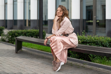 Young European in a jacket, pleated skirt, coral-colored sneakers resting on a bench in the street near the business center. Horizontal portrait of a beautiful girl.
