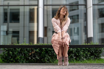 Young European in a jacket, pleated skirt, coral-colored sneakers resting on a bench in the street near the business center. Horizontal portrait of a beautiful girl.