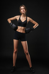 Fototapeta na wymiar Full length portrait of slim caucasian woman in sportswear posing at camera with boxing gloves during workout in gym
