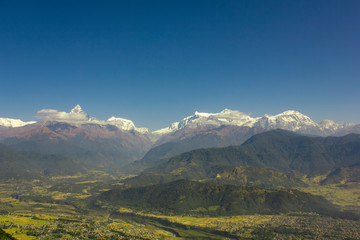 village with a river in a green mountain valley against the wooded slopes and snowy peaks of Annapurn in white clouds under a blue sky