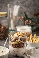 Dessert in a glass of yogurt, granola, banana and chocolate with cookies, cappuccino coffee. Breakfast at the table on a gray background, close-up.Life style composition in dark style.