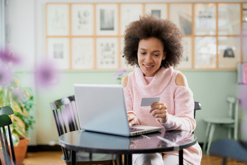 Beautiful mixed race woman in pink turtleneck sweater sitting in pastry shop and using laptop for on line shopping.