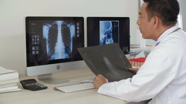 Back view of middle-aged male Asian doctor sitting at desk in his office, looking at computer screens with x-ray images on them and comparing them with x-ray pic in his hands