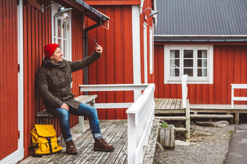 Traveler man with a yellow backpack wearing sits near the wooden red colored house and taking self-portrait a photo with a smartphone. Travel lifestyle concept.