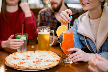 Cheerful friends having fun eating pizza and drinking beer and juice in pizzeria. Cropped image of hands holding glasses with drinks