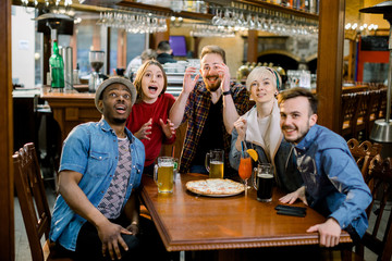 Excited diverse friends having fun watching sports game online video on tv drinking beer eating pizza together, five multiracial people having fun cheering favorite sport team