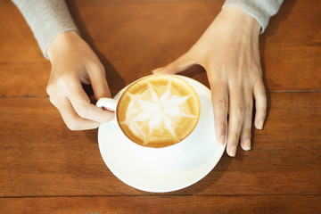 Female hands holding cup of coffee with cobweb shape foam over ready to drink on rustic wooden table, top view. filter tone vintage