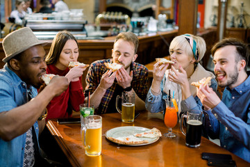 Young five people eating pizza and laughing while sitting in a fast food restaurant. Group of friends enjoying while having food and drinks at cafe.