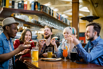 Young five people eating pizza and laughing while sitting in a fast food restaurant. Group of friends enjoying while having food and drinks at cafe.
