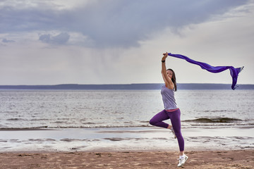 Young slim brunette woman jumping while jogging while holding a blue scarf in her hands. A woman is engaged in gymnastics in the spring morning on the sandy bank of a large river. Cloudy morning.