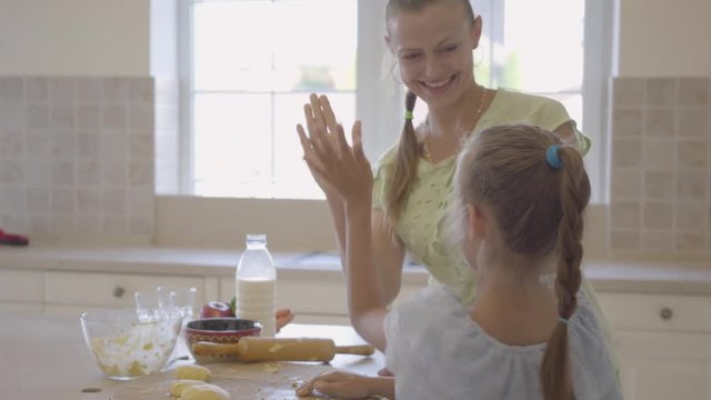 Side view of young woman and her little daughter cooking cakes at home in the kitchen. The girl finishing work and high five with mother. Mom and child spending time together. Real happy family.