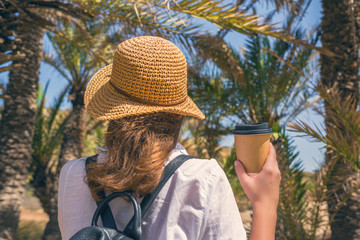 Back view on the Woman in the straw hat with paper cup of coffee walking in the palm forest.