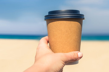 Woman's hand with paper cup of coffee on the beach and sea background.