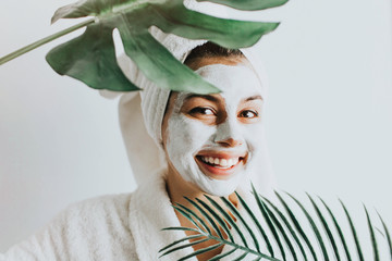Young beautiful woman in face mask of therapeutic white mud and tropical leaves. Spa treatment,...