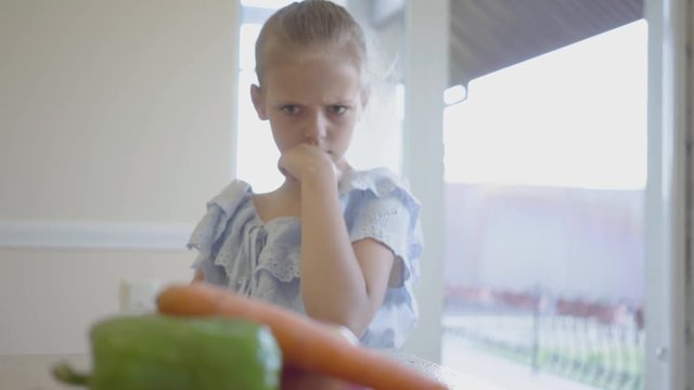 Unhappy angry little girl in front of plate with vegetables in the kitchen. The child pushes away a plate of tasteless food. The kid refusing to eat pepper and carrot