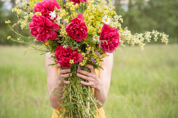 A girl in a dress holds a large bouquet with peonies and wild flowers on a green meadow in summer