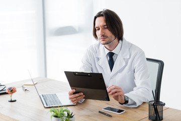 handsome doctor with clipboard in hand sitting in office at desk
