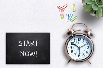 Alarm clock next to slate plate with text "Start Now!" on a white table