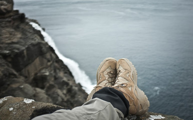 First person view on tourist legs (beige trekking shoes, grey and black membrane clothes) crossed; long waterfall, grey stone cliff and dark blue ocean in the background