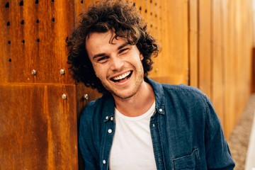 Close up portrait of cheerful young man with curly hair, smiling and looking directly at camera, posing at modern building wall outdoors. Portrait of happy smart student male. People, emotion concept