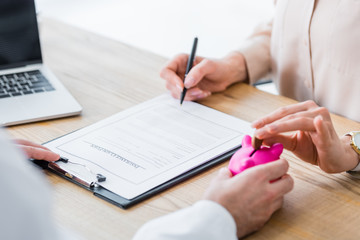 partial view of doctor holding piggy bank in hand while woman holding coin and signing document