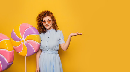 curly red-haired positive woman with lollipops pointing hands into empty yellow background
