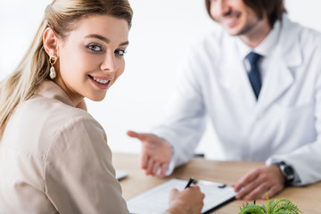selective focus of patient sitting with doctor behind wooden table, signing document and looking at camera