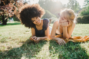 Two young women are lying on the grass in a park while they are joking - Back light portrait of millennials having a summer day at sunset in the city - 275284537