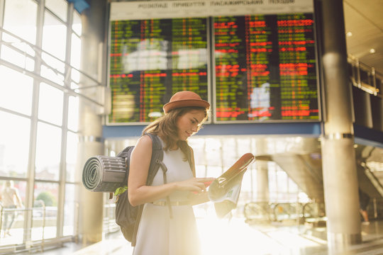 theme travel and transportation. Beautiful young caucasian woman in dress and backpack standing inside train station terminal looking at electronic scoreboard holding phone, map paper hand navigation