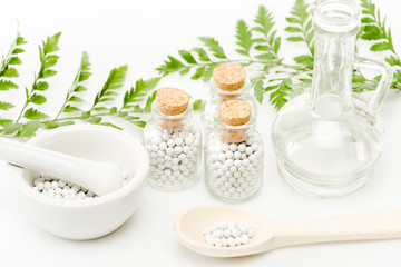 Fototapeta na wymiar glass bottles with small pills near mortar and pestle, wooden spoon, jar and green leaves on white