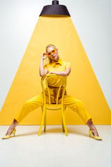 blonde woman touching sunglasses and sitting on chair on white and yellow