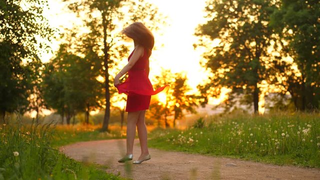 Happy young adult woman dance in park, stay on place and spin around, light red dress fly in air. Beautiful golden sun light at evening, green nature on background