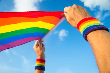 Hands with rainbow color wristbands waving gay pride flag backlit in the wind against a vibrant blue sky
