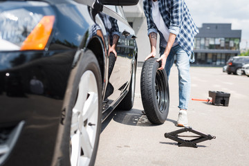 cropped view of man in casual clothes rolling new wheel and fixing broken auto, car insurance concept