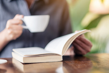 Closeup image of a woman reading a book while drinking coffee on wooden table