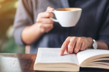 Closeup image of a woman pointing and reading a book while drinking coffee on wooden table