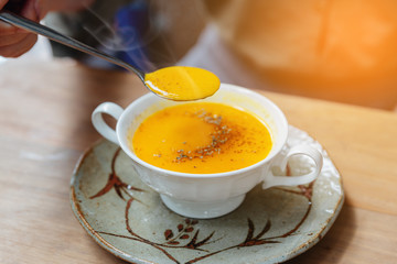 Hand scooping Classic Pureed Pumpkin Soup with steam and topping with pepper. Served in ceramic cup on wooden table.