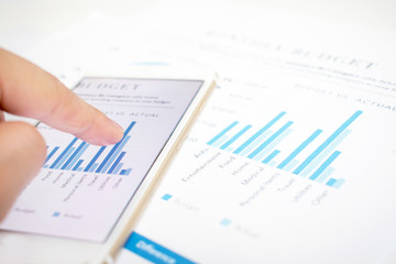 Close-up pictures of businessmen pointing to the smartphone screen showing some graphs. Modern white desk with financial documents.