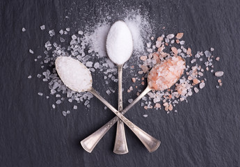 Various kinds of salt in silver spoons on black stone background, top view