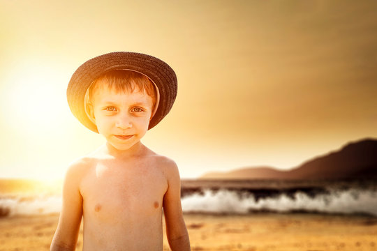 Small young boy on beach with summer hat and blurreed landscape of beach at sunset time. Copy space and natural moment of childhood happiness and family vacation concept.