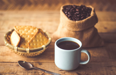 Black coffee In the cup sky blue color have roasted coffee beans are In a cloth bag sack butter cracker In the basket weave bamboo, and still have stainless steel spoon all placed on a wooden table.