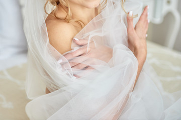 Portrait of beautiful bride in lingerie and veil with crossed hands sitting on bed in room, copy...
