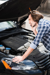 frustrated man talking on smartphone while standing near broken auto with open trunk, car insurance concept