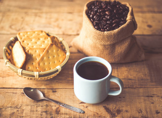 Black coffee In the cup sky blue color have roasted coffee beans are In a cloth bag sack butter cracker In the basket weave bamboo, and still have stainless steel spoon all placed on a wooden table.