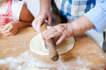 Hands of grandmother roll out dough with rolling pin and flour on wooden table. Family is cooking pastries in kitchen. Senior woman and child prepare homemade pie or pizza