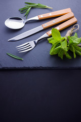 Cutlery set. Knife, spoon, fork on slate stone. Сutlery with rosemary and mint. Black background. Copy space. Top view